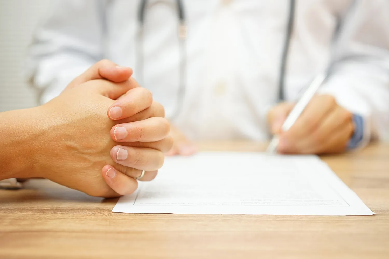 Patient with clasped hands across a desk from a doctor writing on a report.