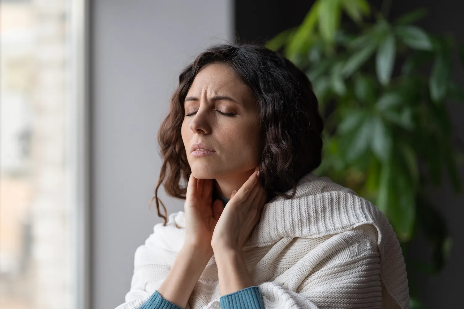 Woman wrapped in a sweater holding her sore neck with eyes closed.