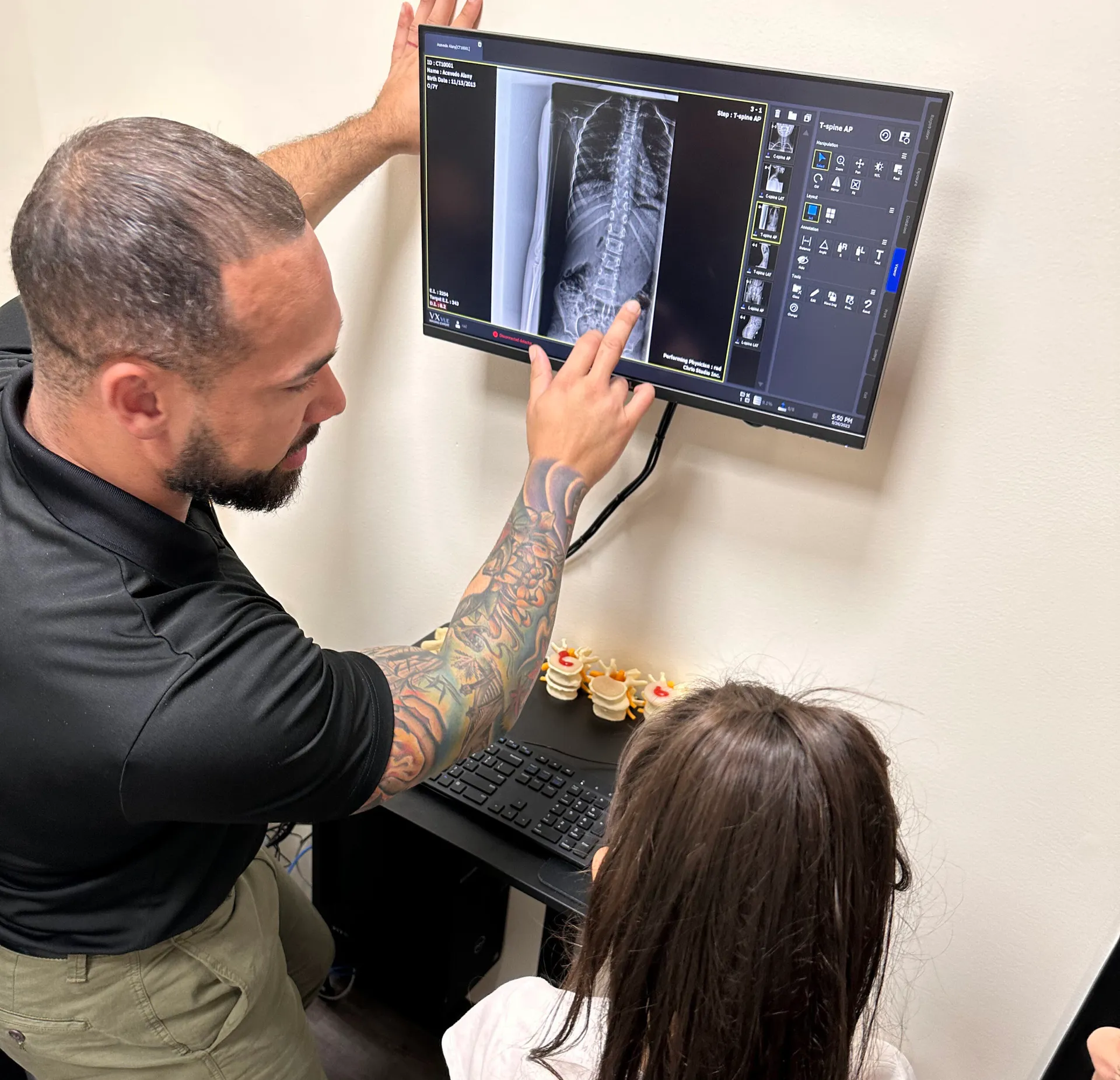 Dr. Junior pointing to a spinal X-ray on a wall monitor while a young girl watches.