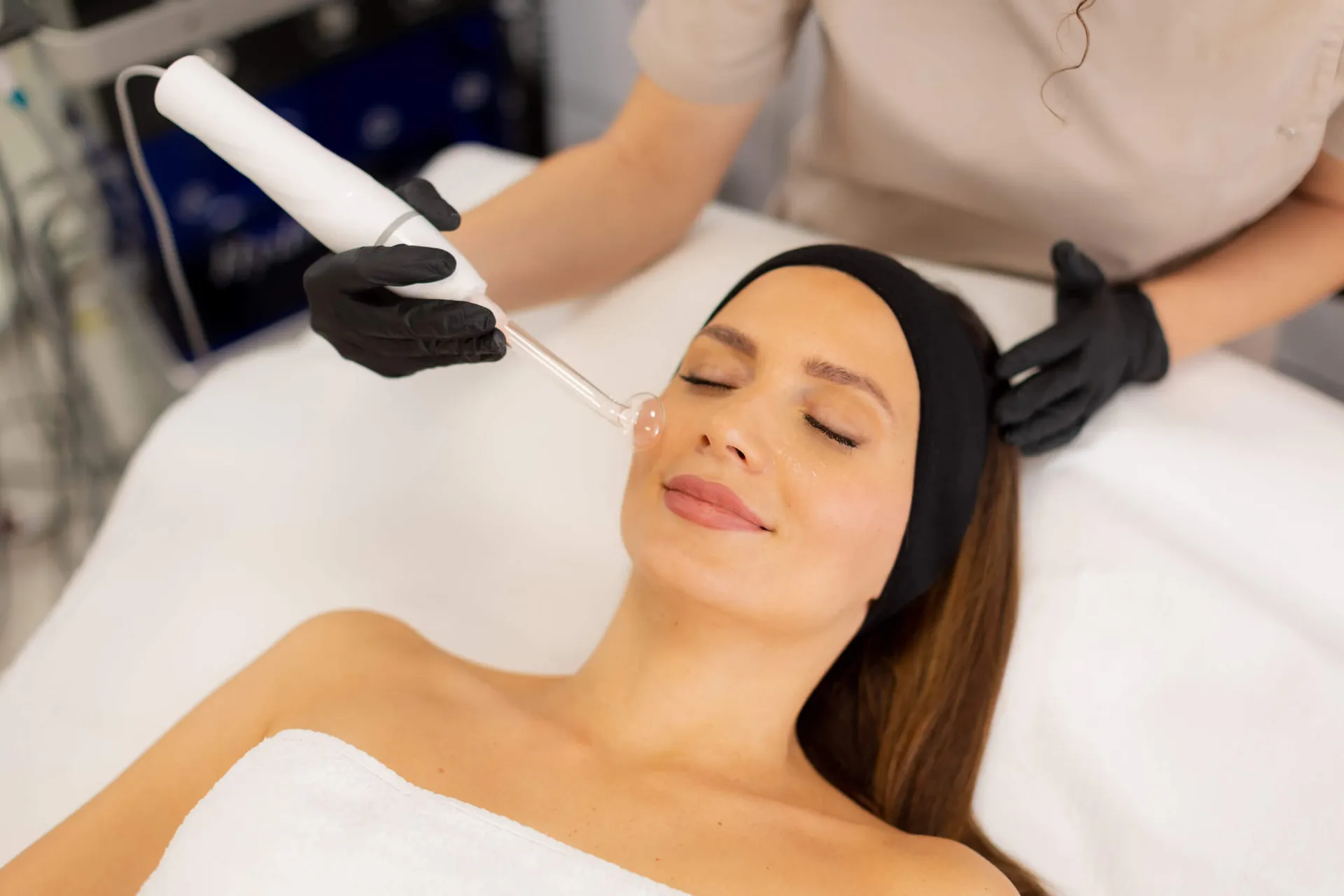 Woman relaxing on a spa bed while a gloved technician applies a handheld wand to her face.