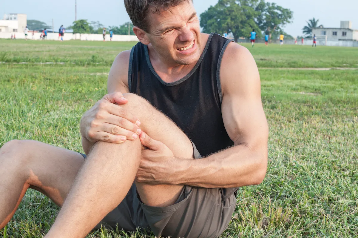 Athlete sitting on a grass field grimacing and holding his injured knee.