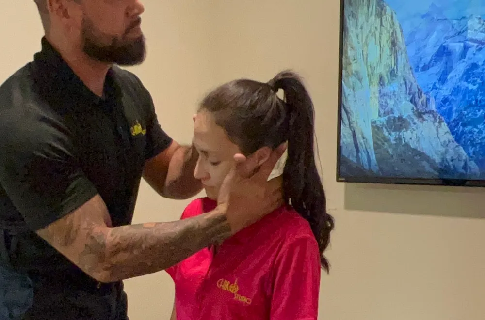 Chiropractor cradling the head of a seated young woman in a pink shirt to check her neck.