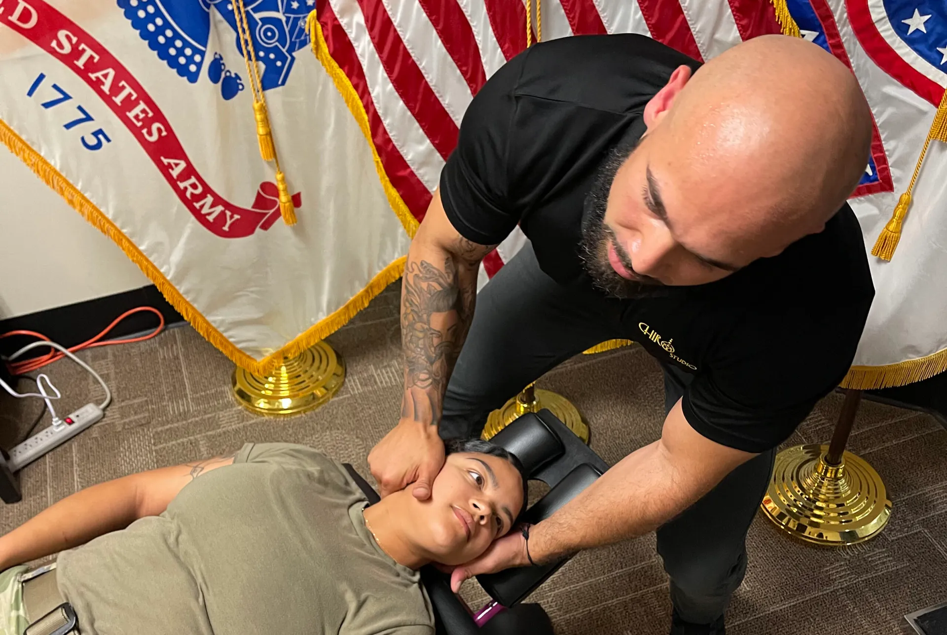 Dr. Junior adjusting the neck of a soldier lying face up in front of U.S. Army and American flags.