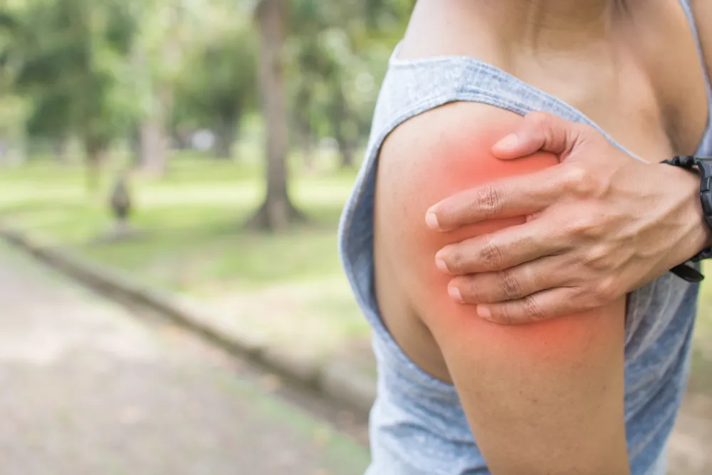 Person gripping their upper arm and shoulder outdoors, with the painful area highlighted in red.